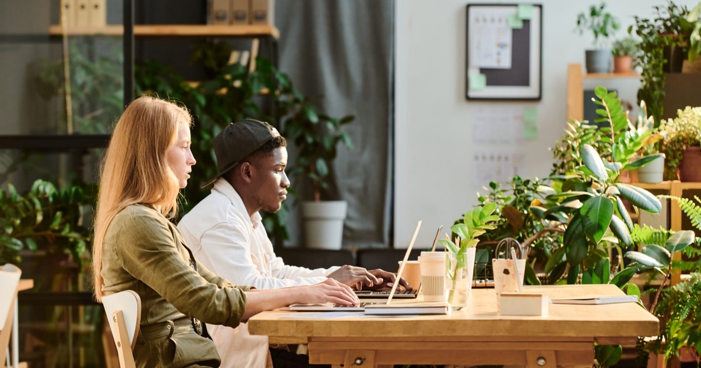 Two individuals seated at a table, each using a laptop, engaged in a collaborative work session.