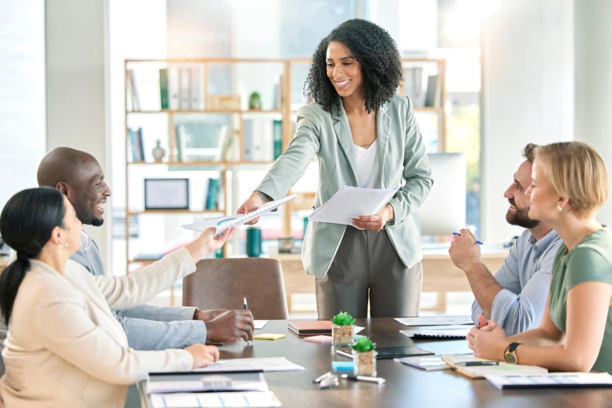 A woman stands at a podium, presenting to a group of attentive colleagues seated in a conference room.