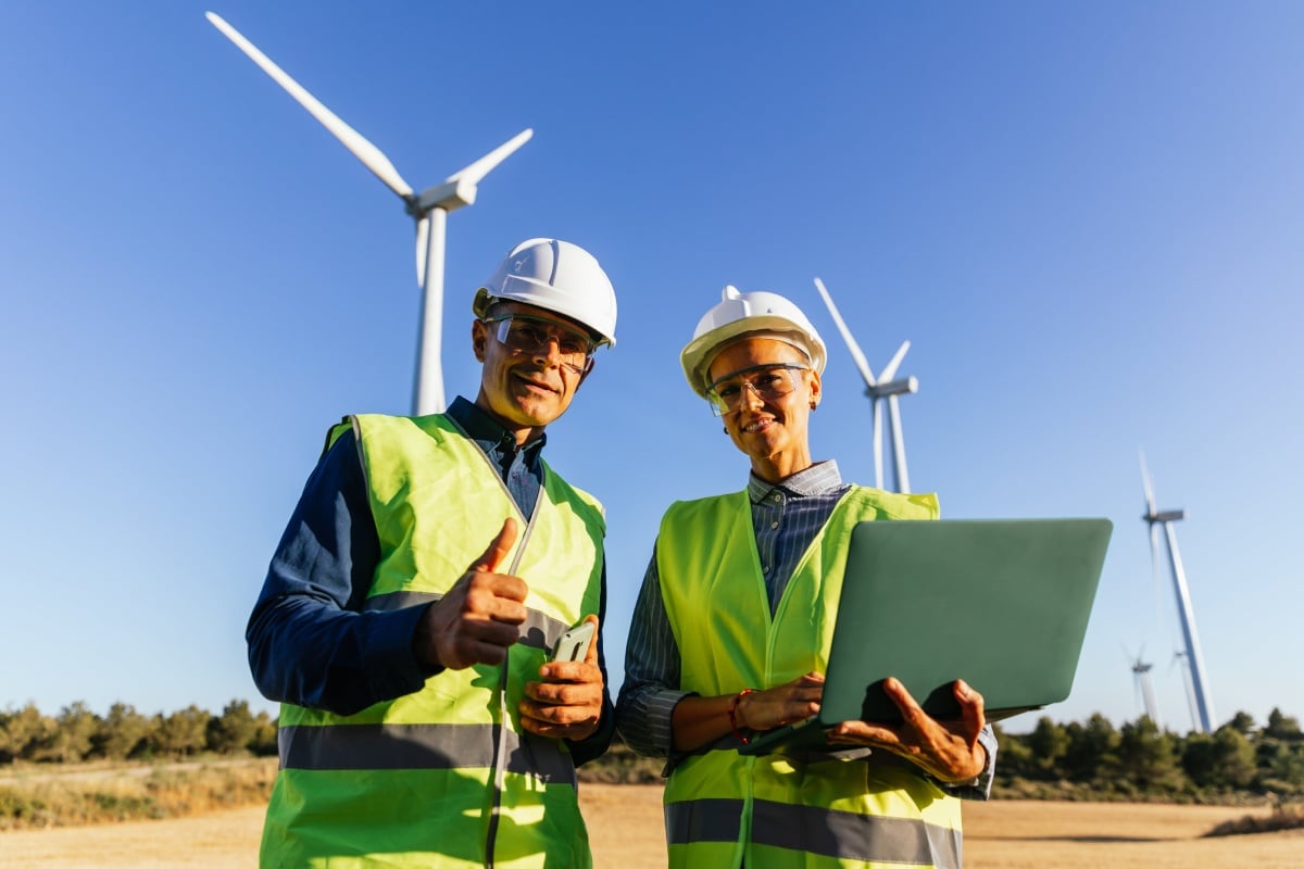 Two men in hard hats and vests examine a laptop, with wind turbines visible in the background.