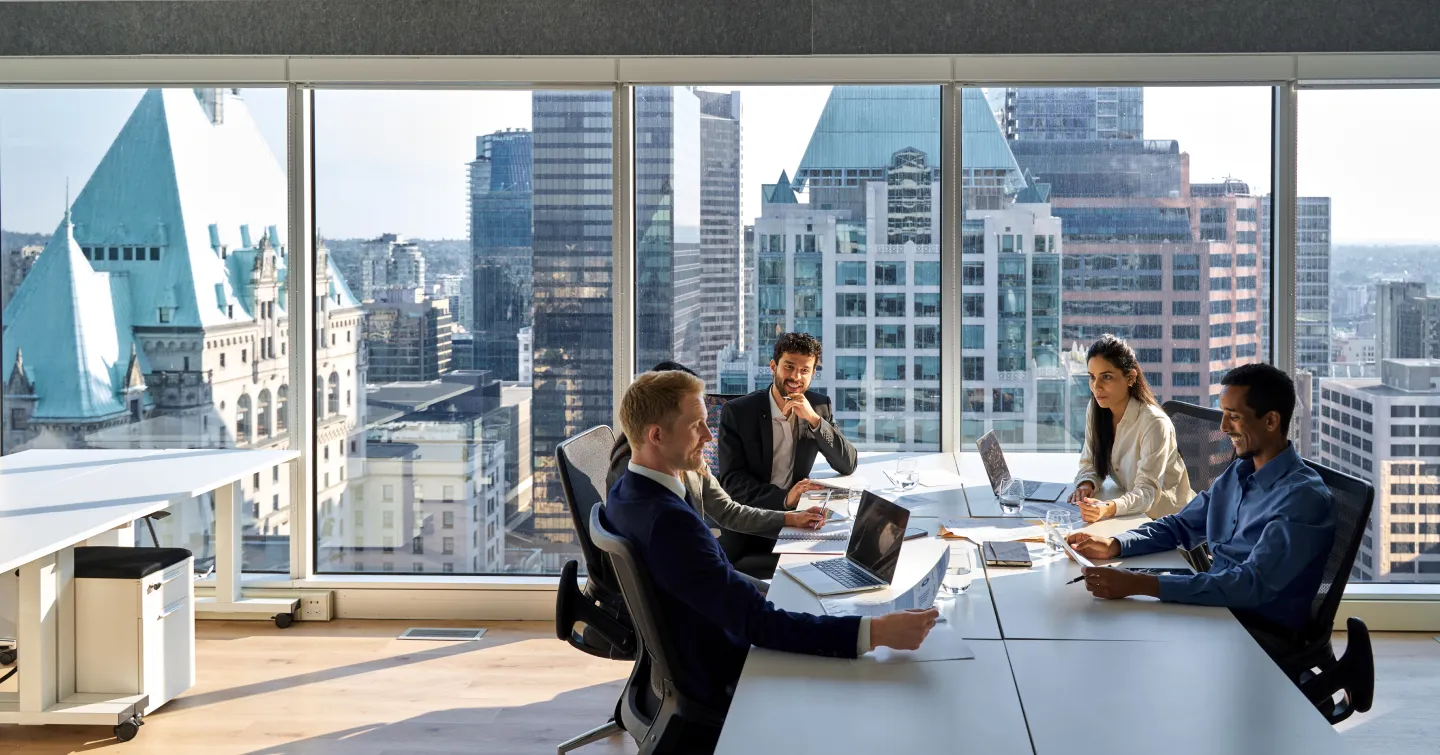 A group of business professionals seated at a table, engaged in discussion, with a large window in the background.