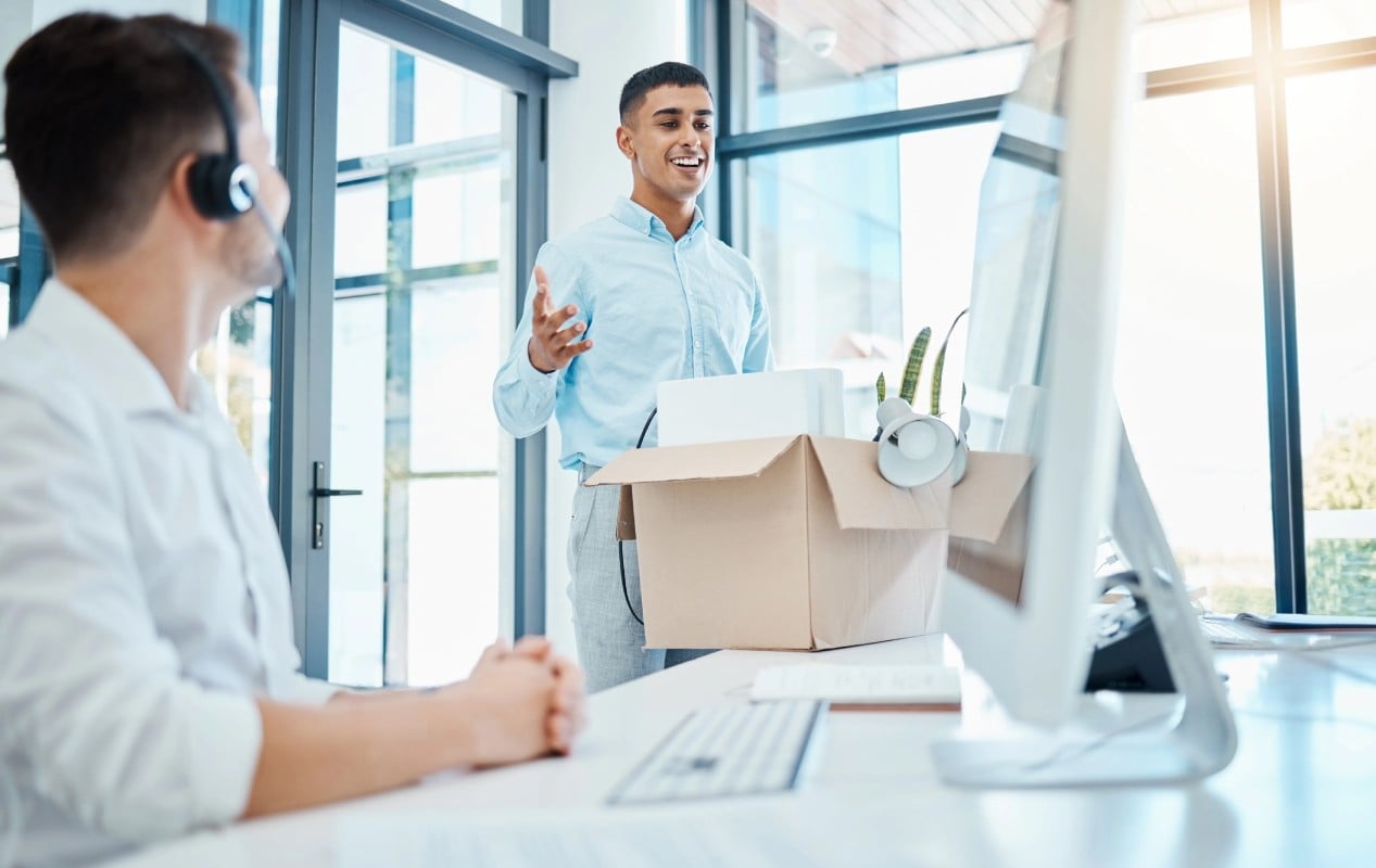 Two men in an office setting, surrounded by boxes on their desks, engaged in a discussion.
