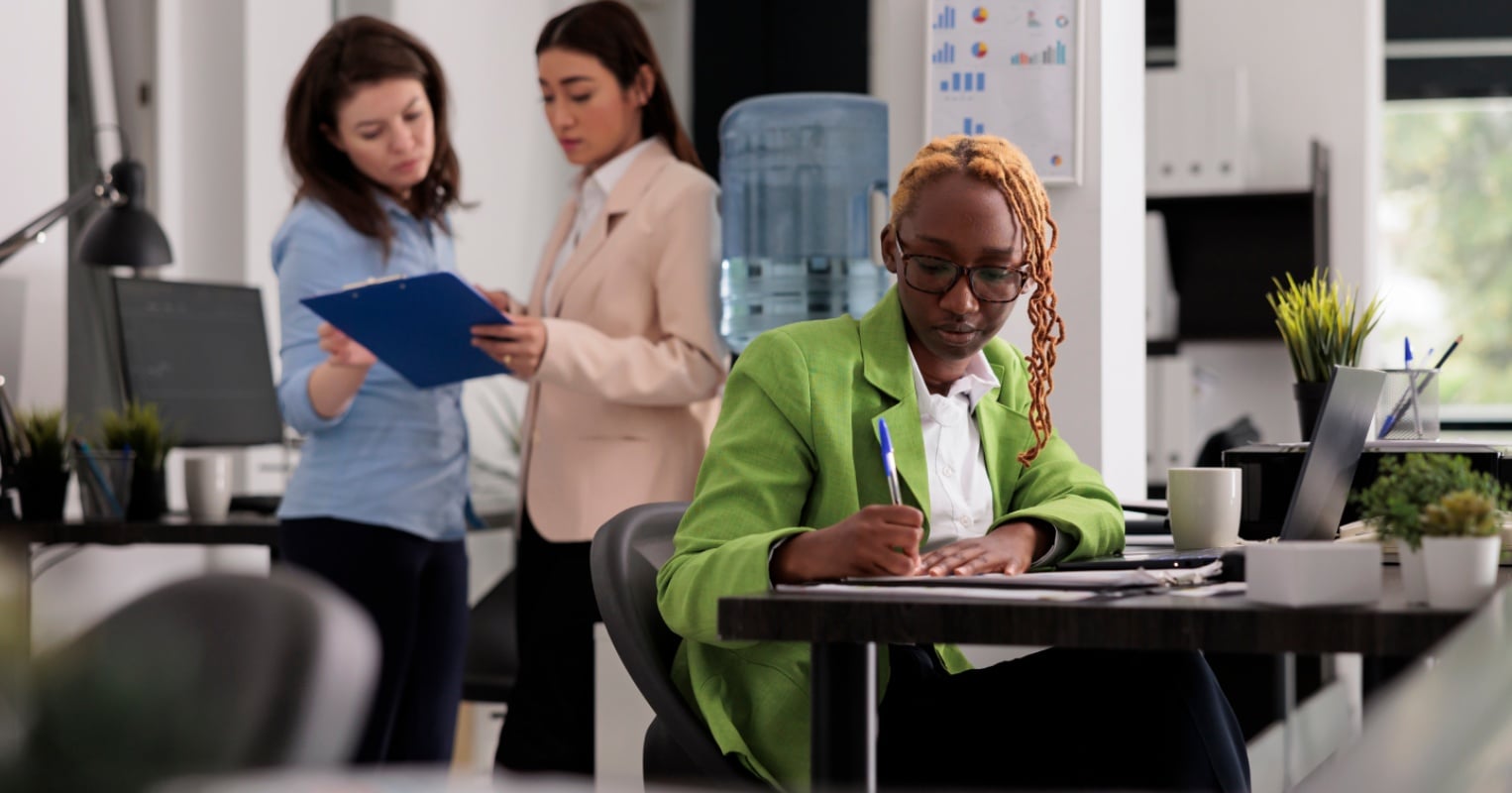 Three professional women working in a modern office; one in green jacket writing at desk, two others reviewing a document.