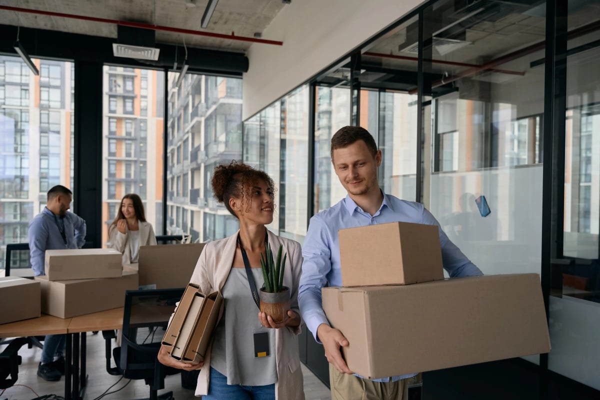 A man and woman carrying cardboard boxes in an office setting, indicating a move or organization of workspace.
