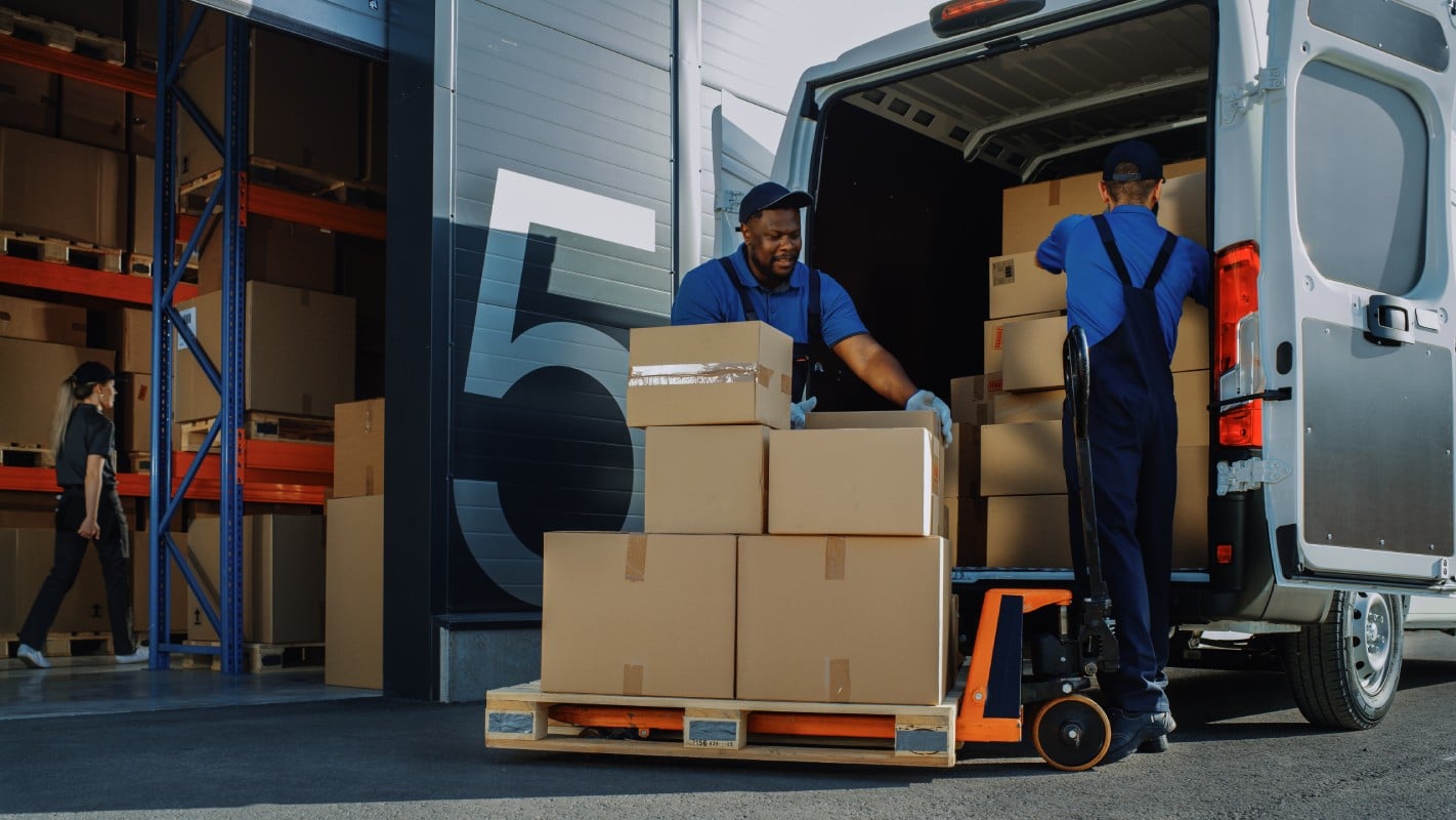 Two men unloading cardboard boxes from a delivery truck in a warehouse setting.