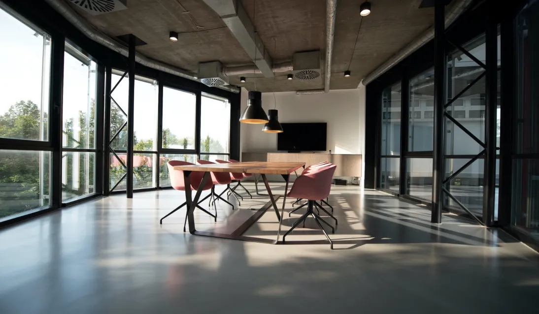 A modern conference room featuring a large table surrounded by pink chairs, large windows allowing natural light, and a minimalist design.