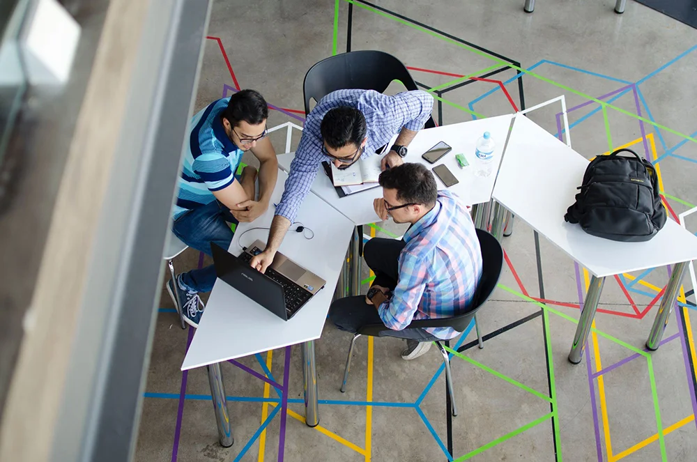 Overhead view of three men collaborating at white, uniquely shaped modular desks on a concrete floor decorated with colorful geometric tape lines.