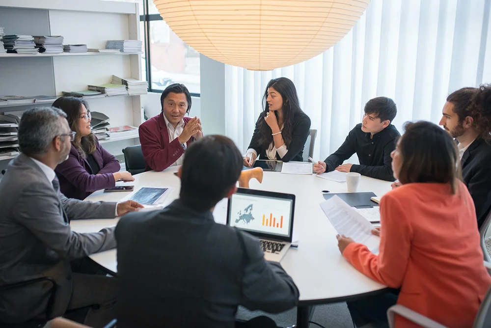 A diverse group of business colleagues sitting around a large round table in a meeting room, listening to a man in a red blazer speak under a large spherical paper lantern.