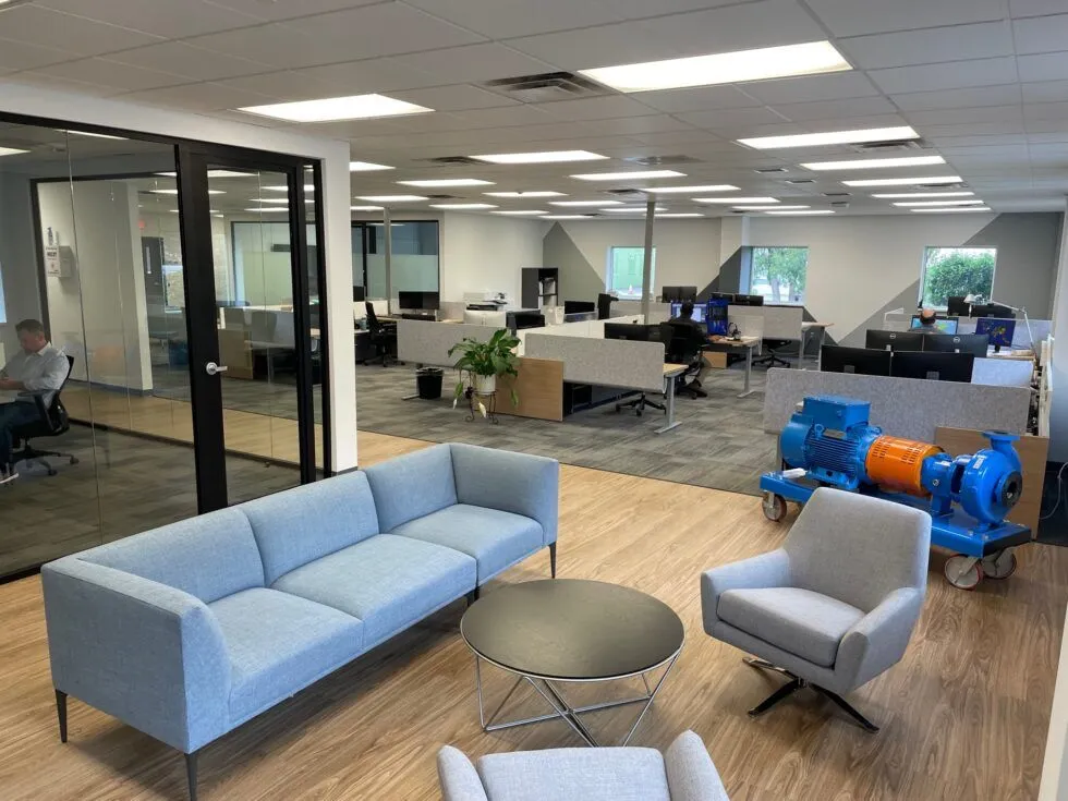An office space with gray couches, a round table, and workstations in the background.