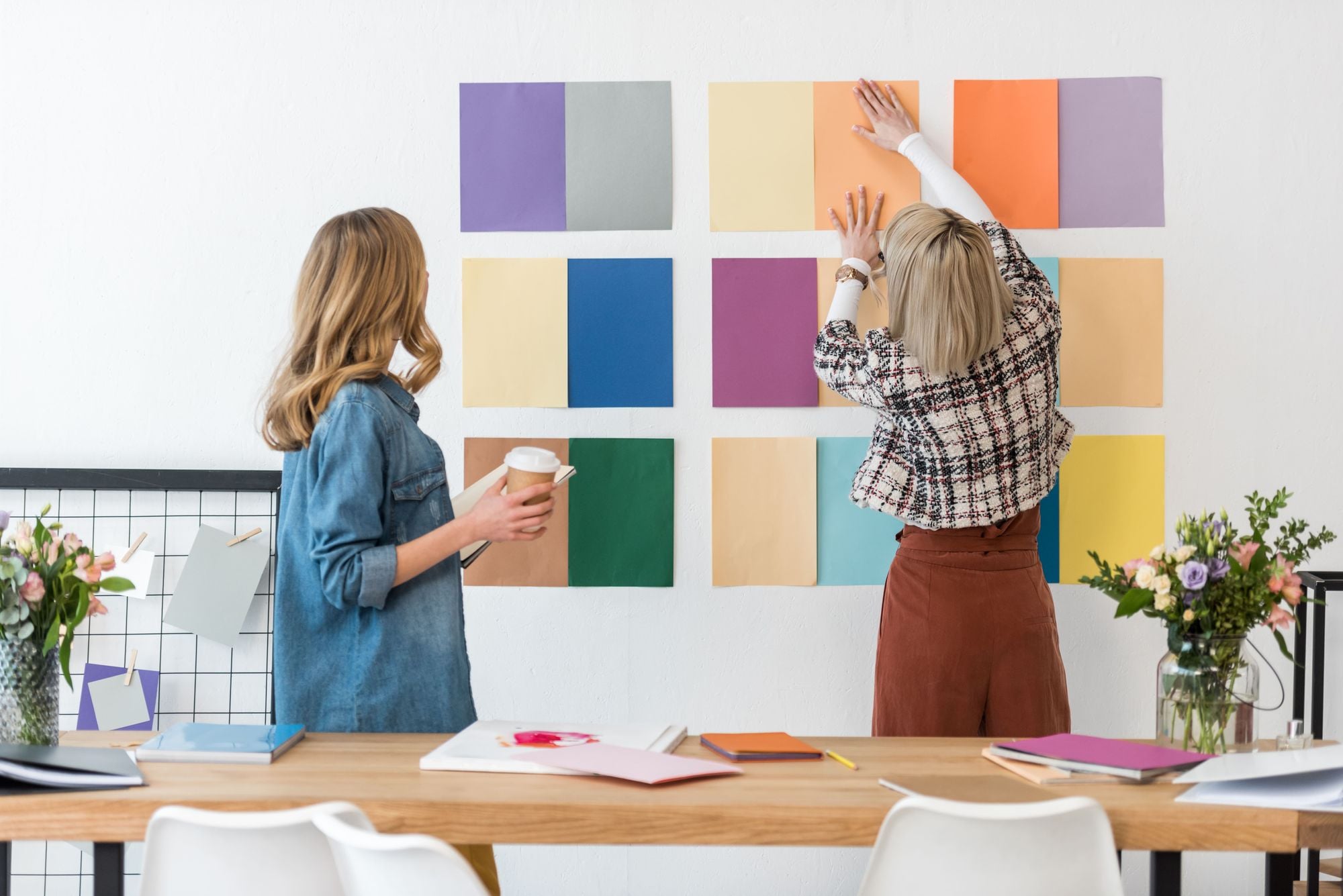 Two women arranging colorful paper samples on a wall in a bright, modern workspace. One woman holds a coffee cup while the other is adjusting the samples. A table with books and flowers is in the foreground.