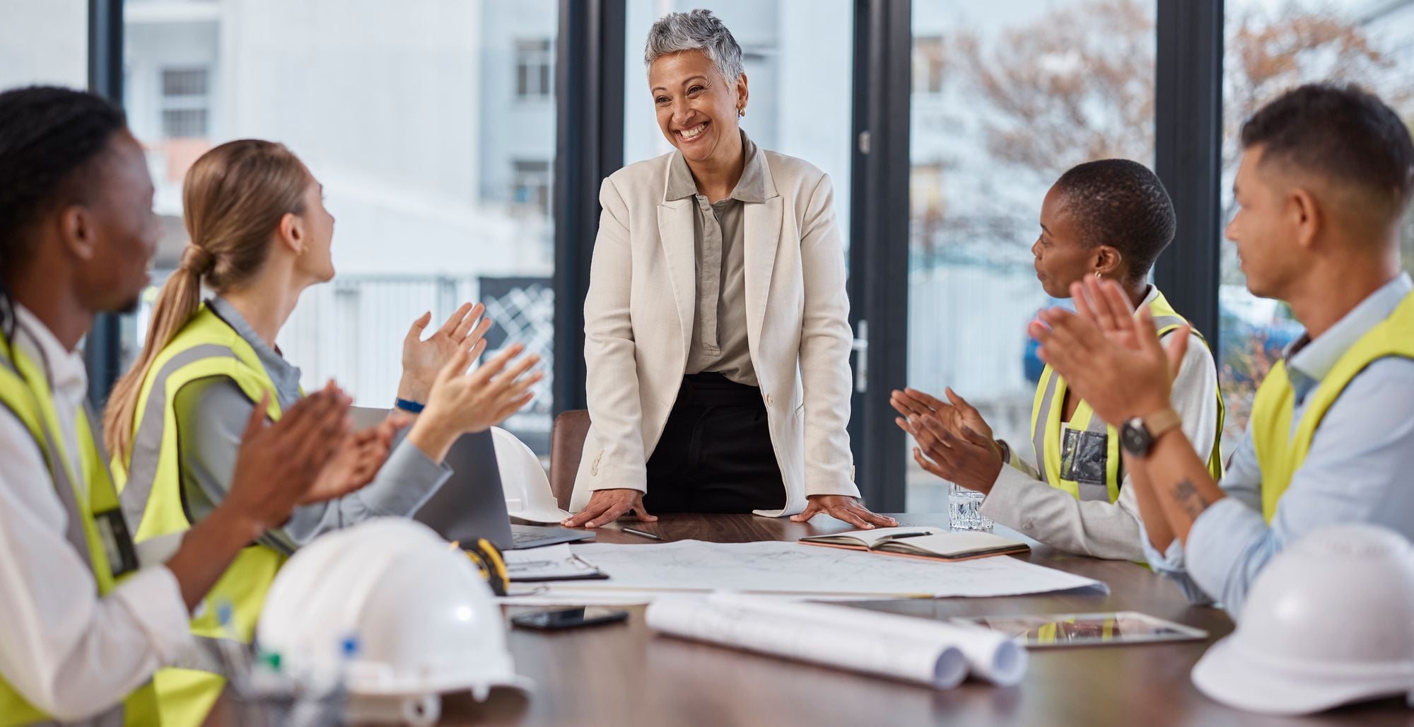 A group of diverse professionals in safety vests engaging in a meeting, with a woman standing at the center smiling, while others are clapping and showing appreciation.