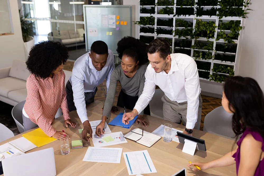 A diverse group of five business professionals leaning over a wooden conference table, collaborating and pointing at documents and charts in an office with a vertical green plant wall in the background.