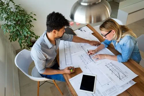 High-angle view of a male and a female architect collaborating on paper building blueprints spread across a wooden table under a metallic dome lamp.