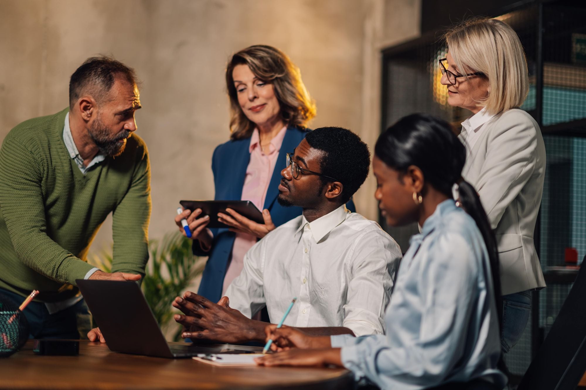 A group of professionals collaborating in an office setting, with two men and two women engaged in a discussion around a laptop.