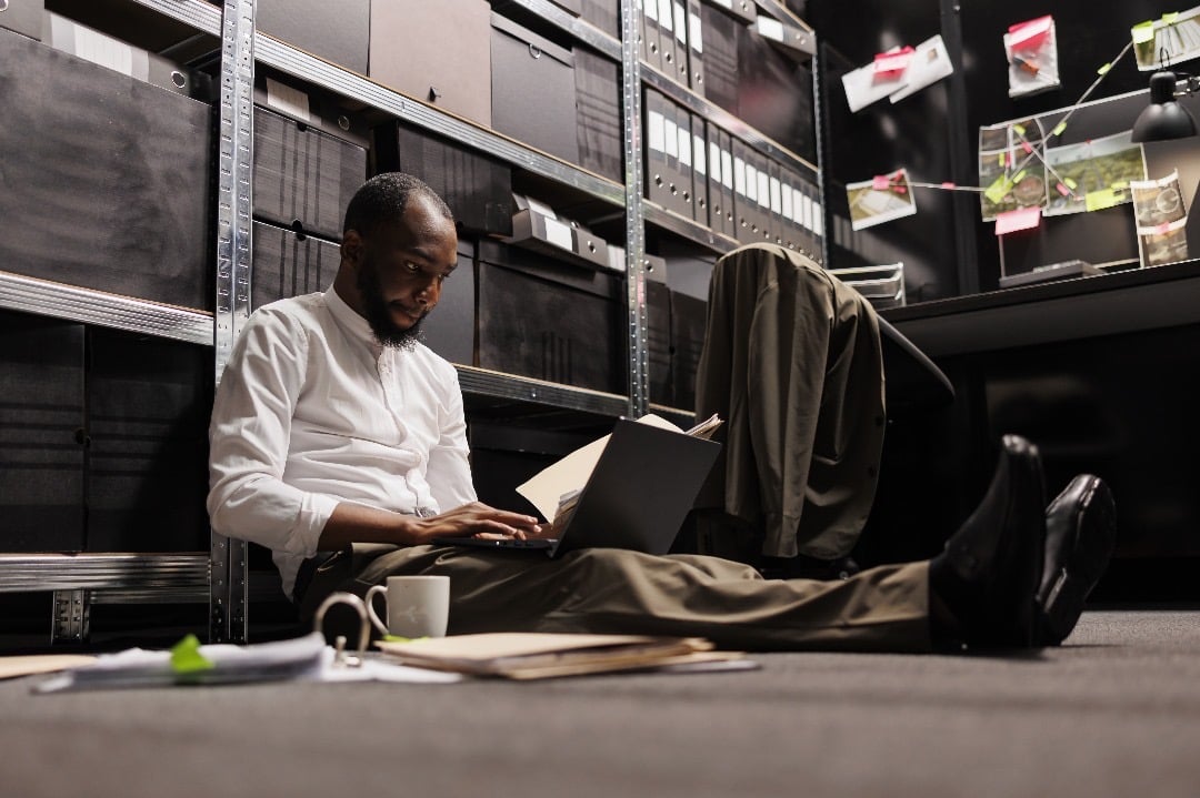 Man sitting on the floor of an office storage room, working on a laptop with files and a coffee cup nearby.