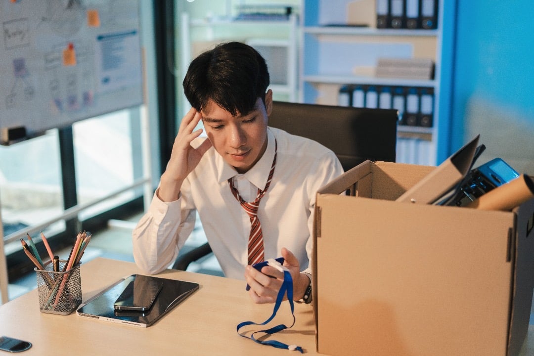 A young man in a white shirt and tie sitting at a desk with a box of belongings, looking thoughtful or stressed.