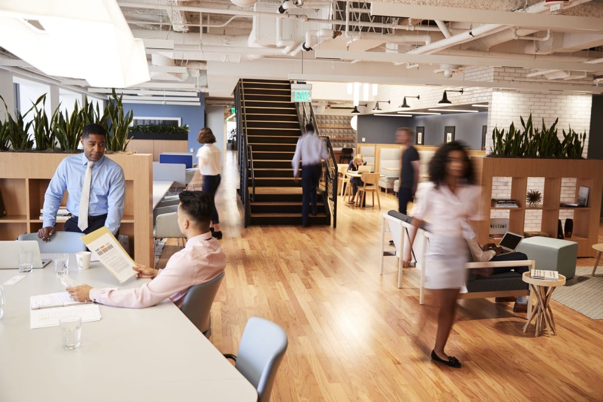 Open office with people working, walking, and discussing documents near stairs and plants in a modern workspace.