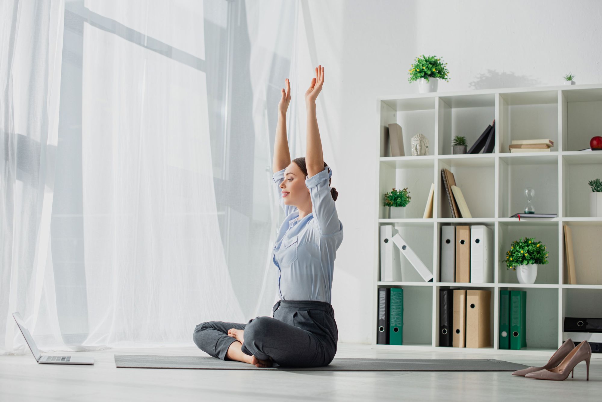 A woman in formal attire practicing yoga in a bright room with a large window and shelves filled with books and decorative plants.