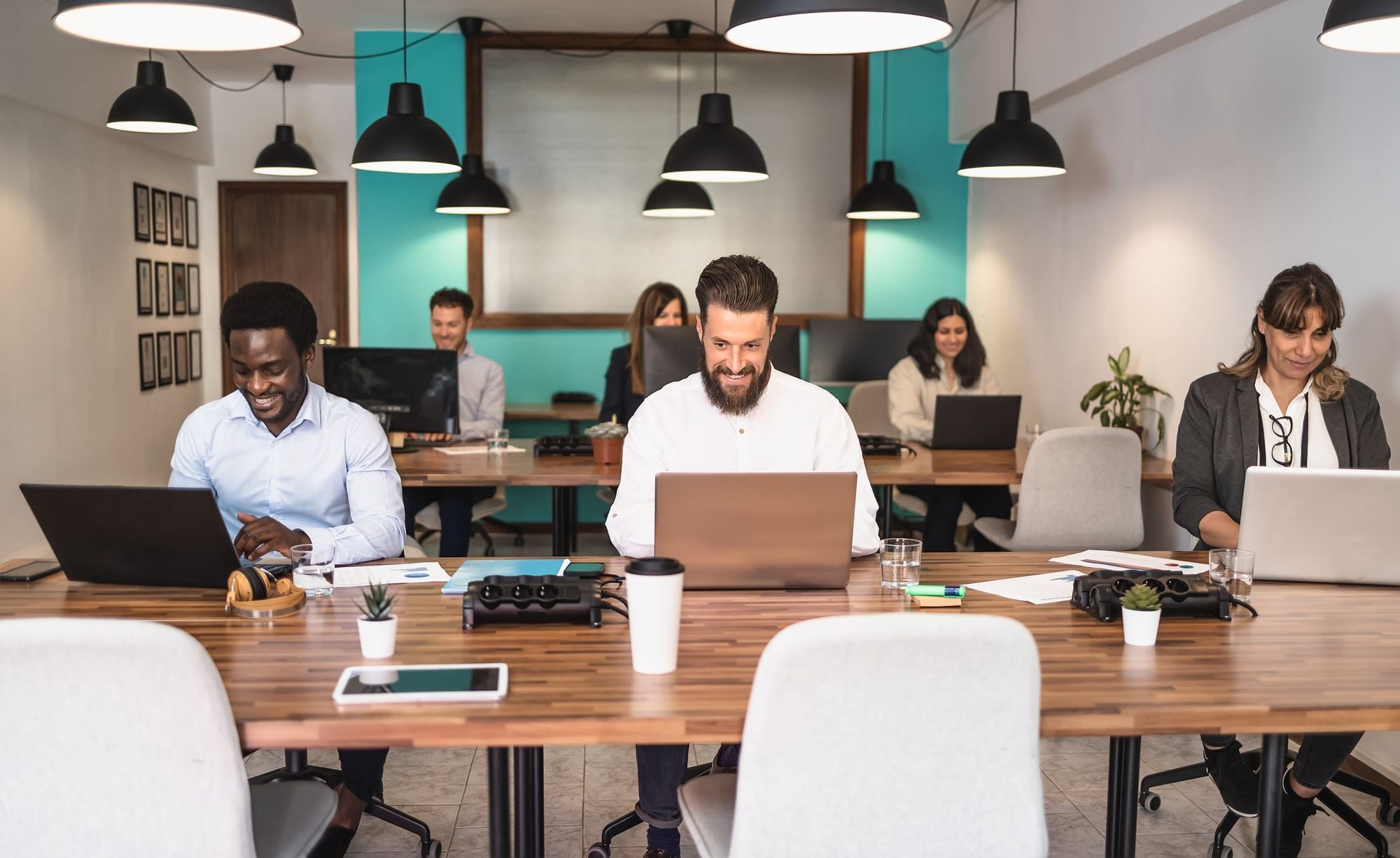 Several people work at laptops in a modern, open-plan office with wooden desks, chairs, and hanging lights.