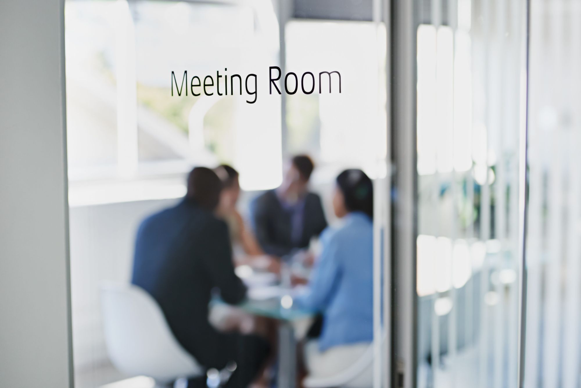Glass door with "Meeting Room" text visible, showing a blurred group of people having a discussion inside.