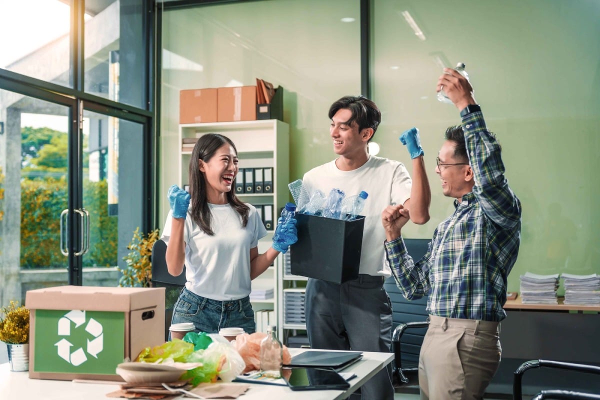 Three people in an office setting celebrating while holding recyclable materials, with a recycling box nearby.