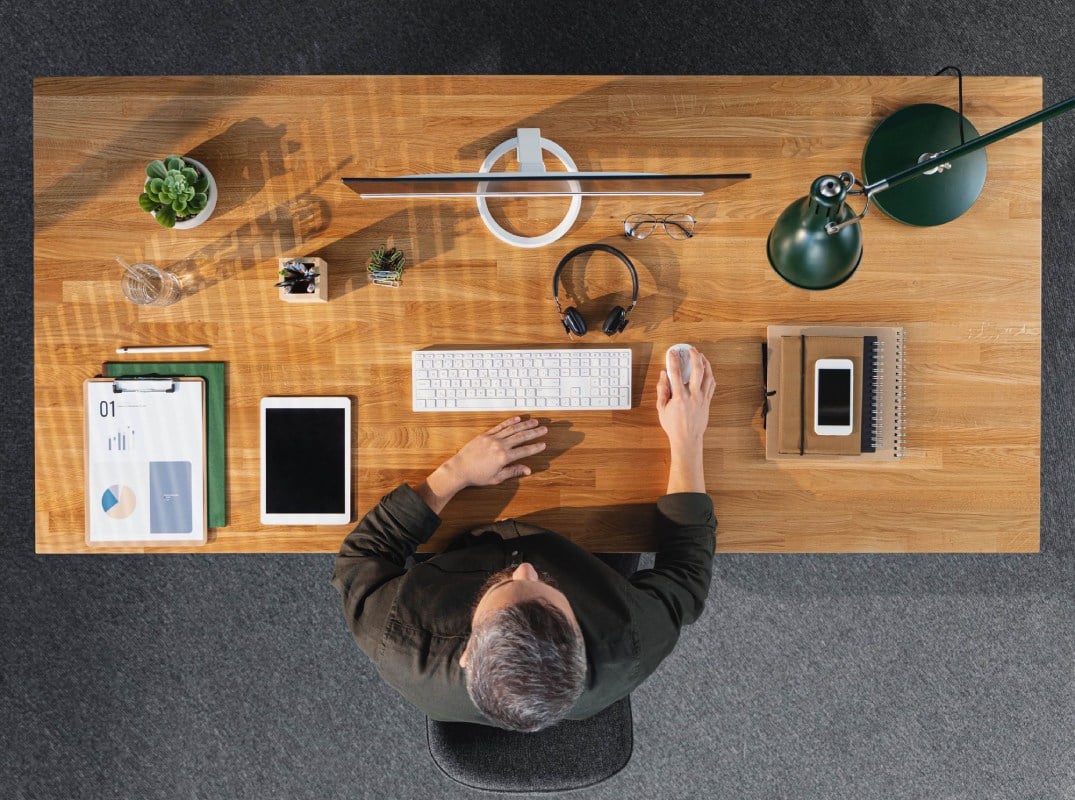 Aerial view of a person sitting at a wooden desk with a computer monitor, keyboard, mouse, tablet, headphones, and various desk items including a notebook and plant.