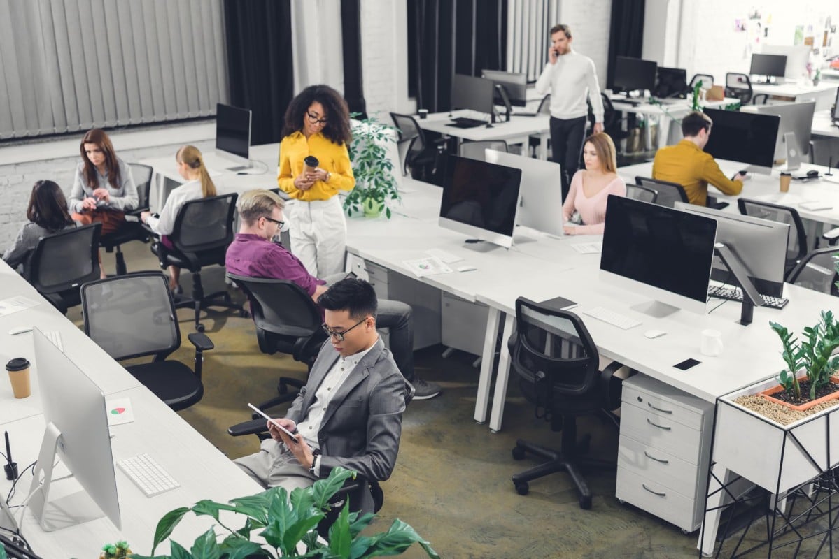 An office environment with several people working at desks, some sitting on chairs using electronic devices, and others standing while engaging in conversation. The workspace features plants and modern furniture.