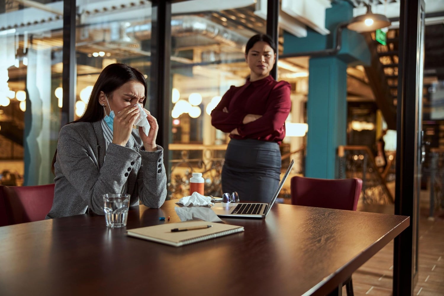 Two women in an office, one sitting and sneezing into a tissue, the other standing with arms crossed, looking concerned.