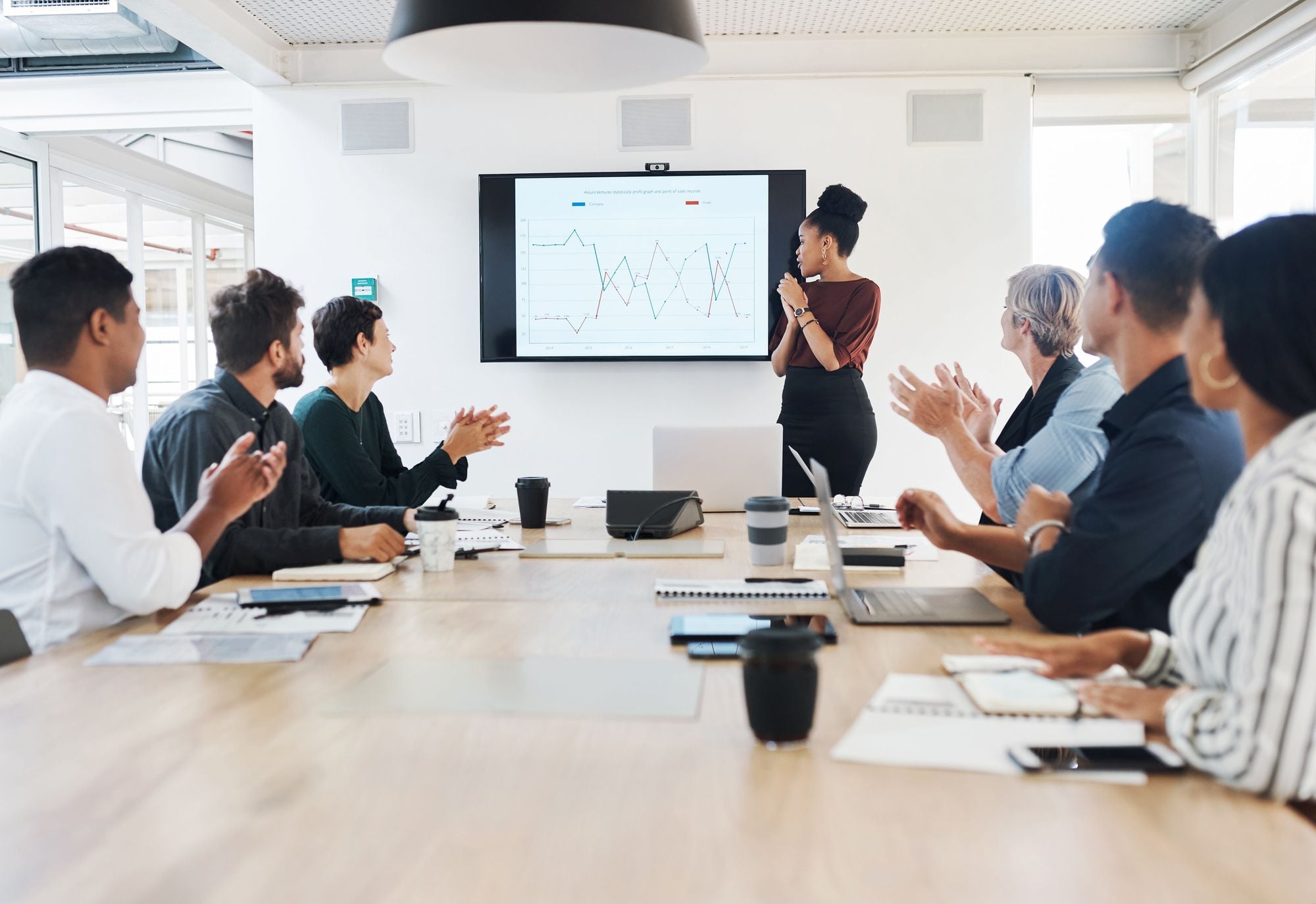 A diverse group of people in a modern conference room, attentively listening to a presenter who is standing in front of a screen displaying a graph.