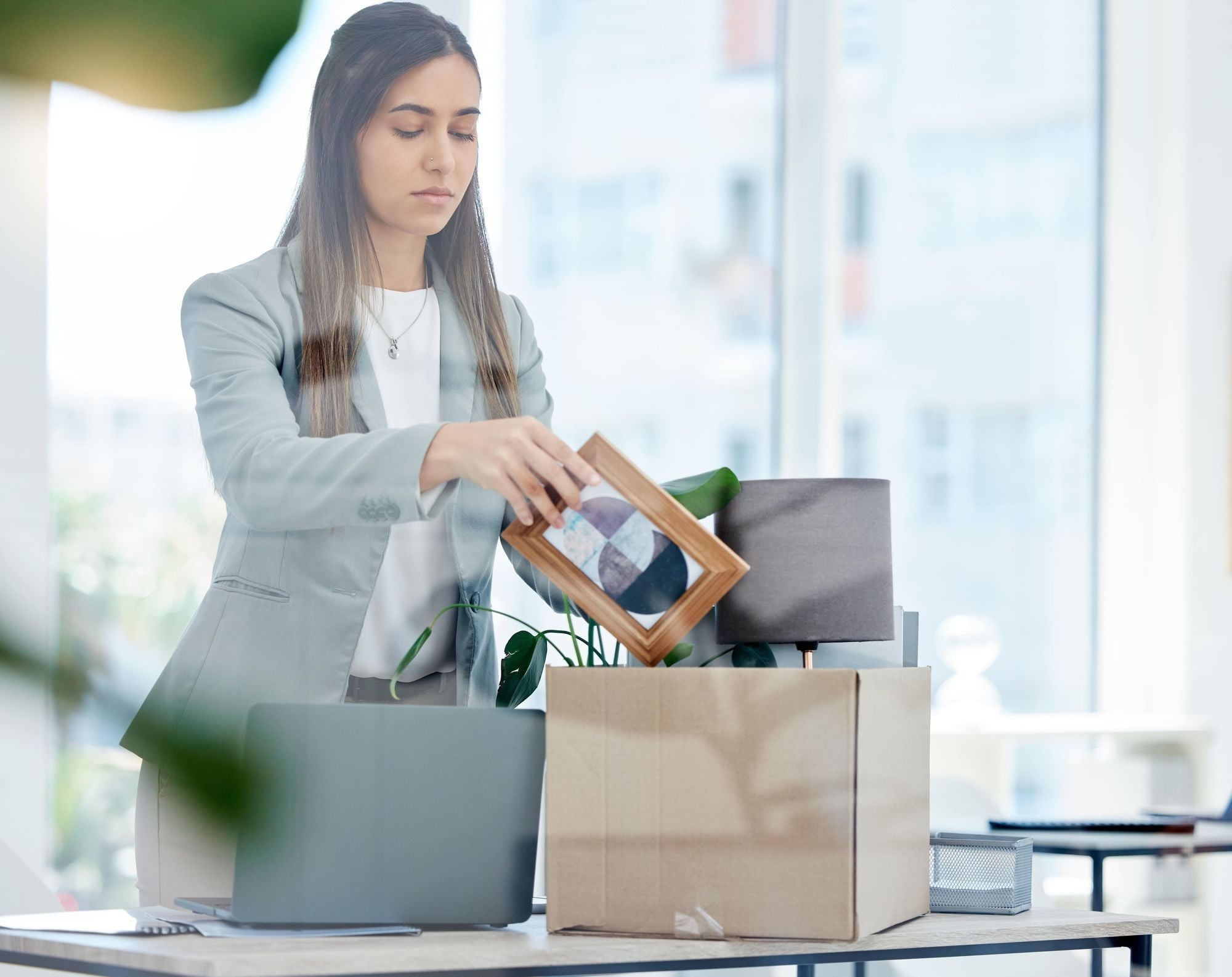 A woman in a professional outfit is packing a box with personal items, including a picture frame, at a desk with a laptop and plants.