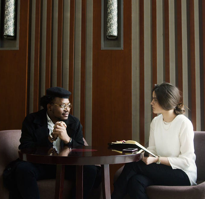 A man in a black beret and glasses sits at a round wooden table, talking with a woman in a white blouse who is holding a notebook, set against a striped wood panel wall.