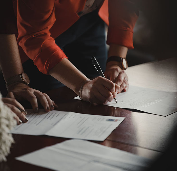 Close-up of a person in an orange blazer signing documents on a table, with another person's hands resting near the paperwork.