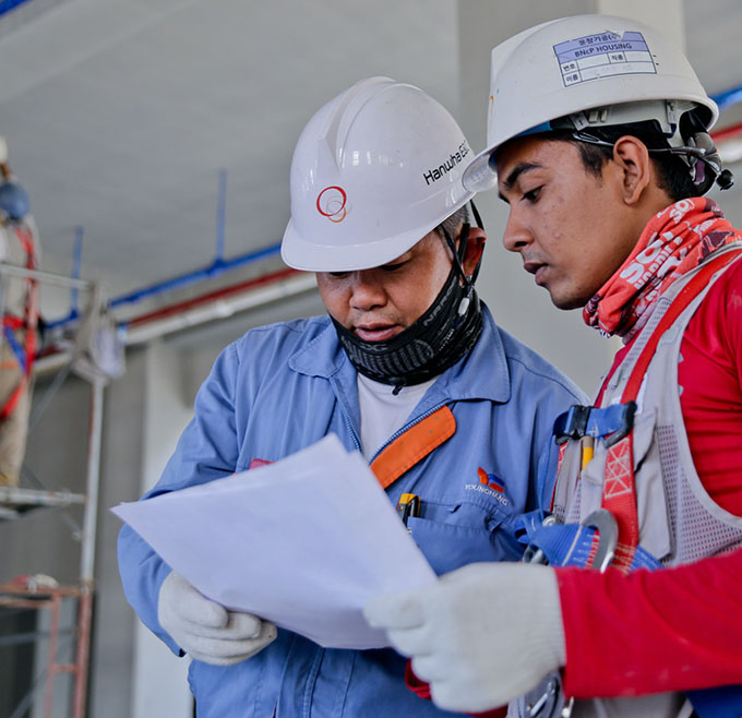 Two construction workers wearing white hard hats, safety gear, and gloves review a white document together at an indoor construction site.