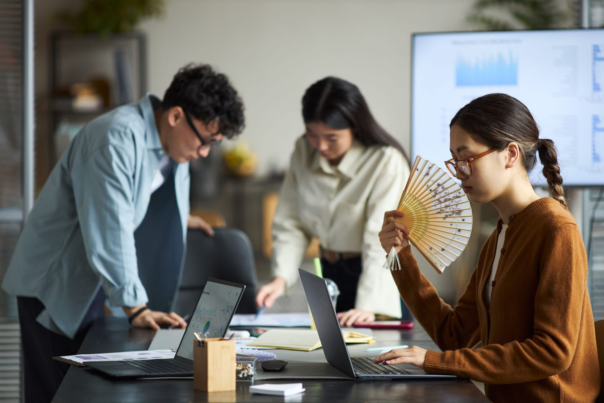 A group of three people working in an office environment, focusing on laptops and documents. One person is holding a fan while seated at the table.