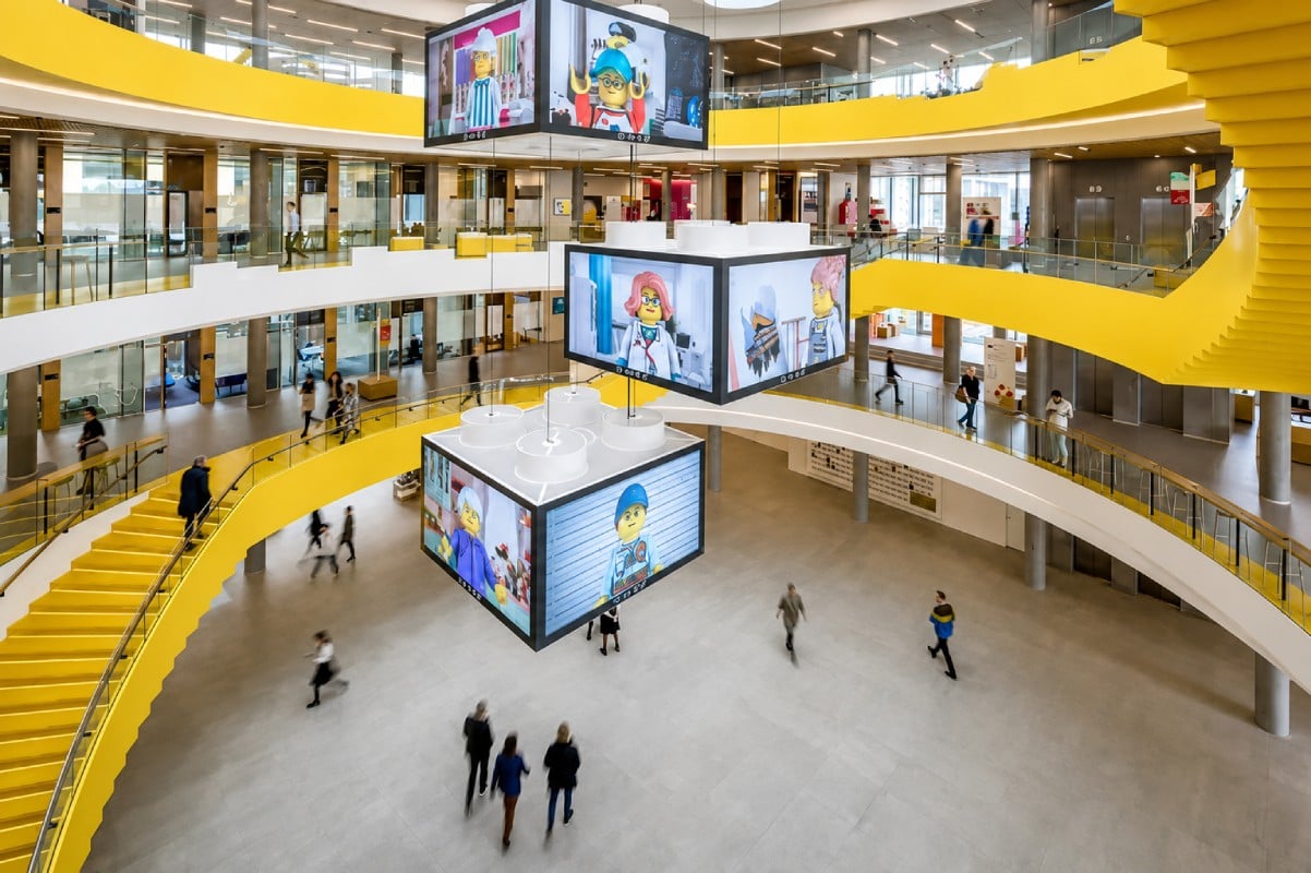 A modern atrium with yellow accents, featuring hanging digital displays and people walking around the open space.