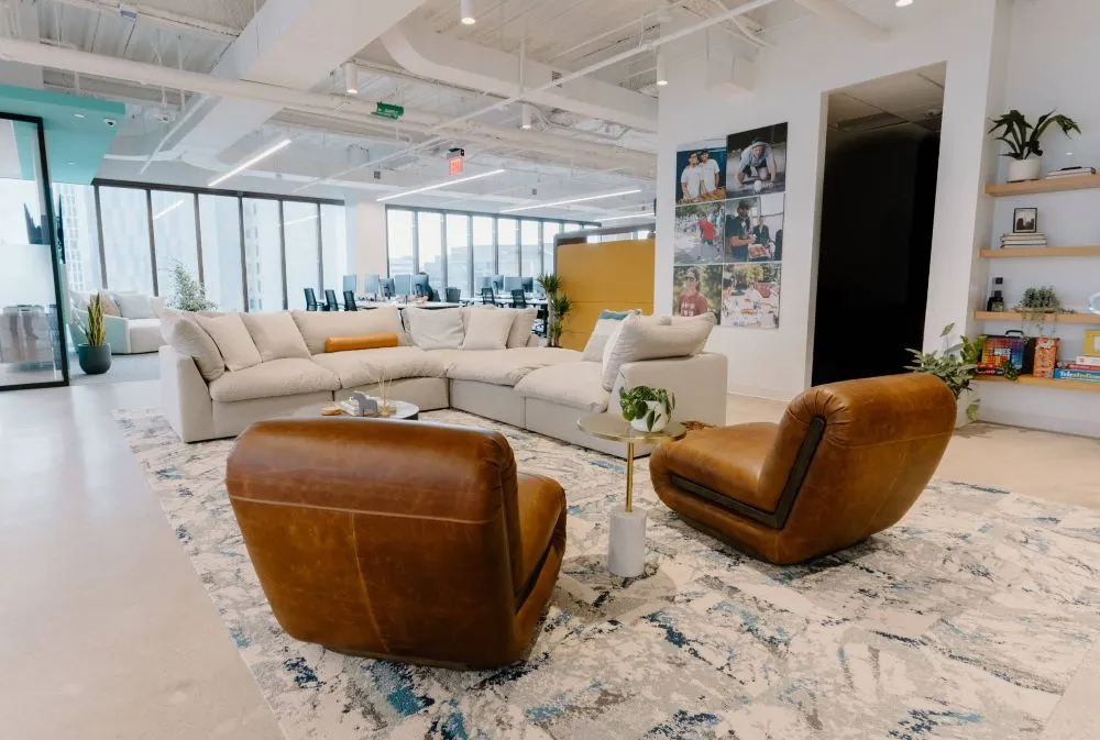 A spacious office collaboration area featuring a large, off-white modular sectional sofa paired with two low brown leather armchairs, arranged around a patterned rug in front of an open workstation area.