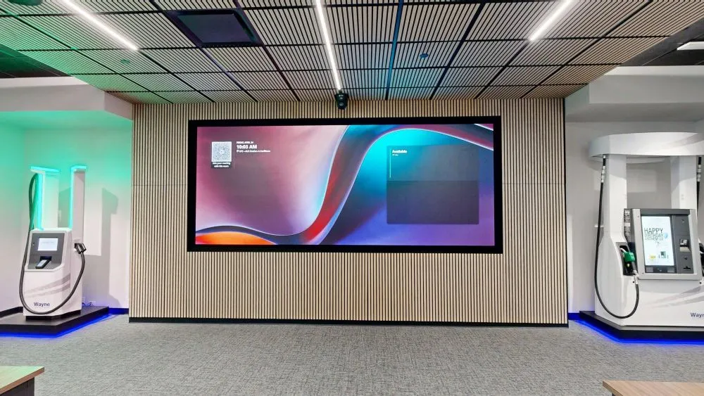 A large digital display screen framed by a wood-slatted feature wall, flanked by model fuel dispensers on each side in a modern showroom setting.