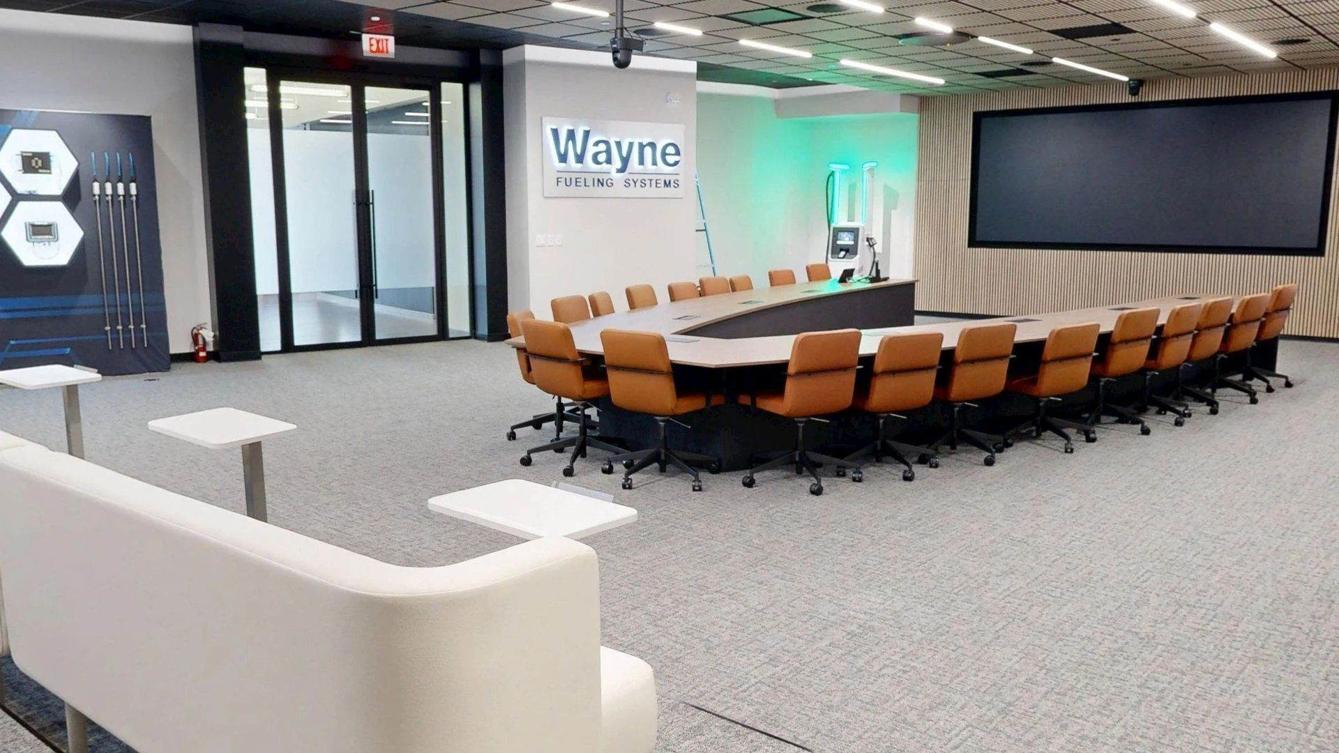 A large presentation room featuring a curved U-shaped conference table lined with tan leather chairs, set in front of a "Wayne FUELING SYSTEMS" wall sign and a large media display screen.
