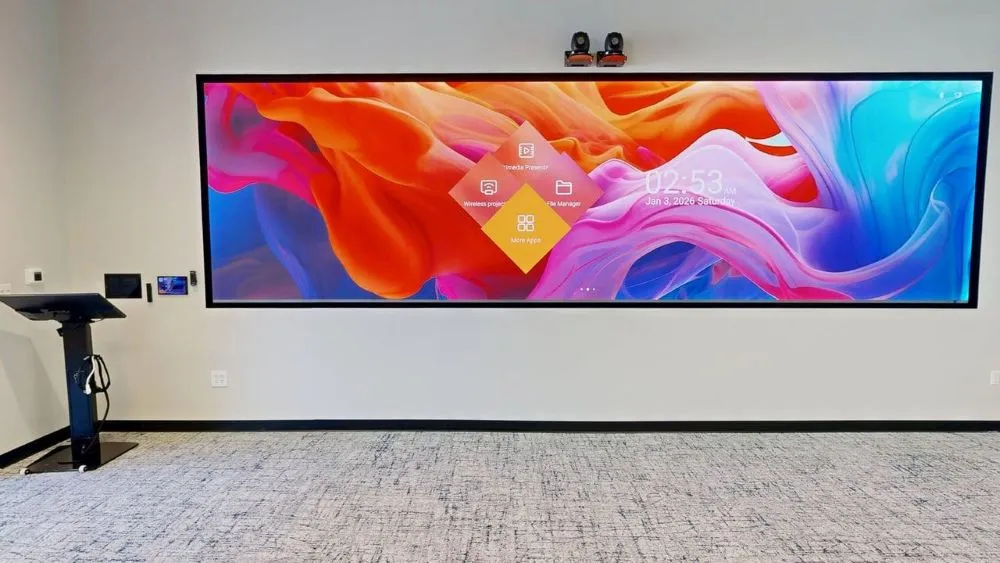 A wide panoramic video display wall showing colorful abstract graphics, mounted above gray carpeted flooring next to a black presentation podium.