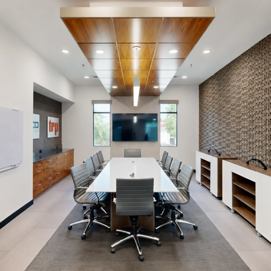 A modern conference room featuring a long white table with dark gray leather office chairs, a wooden ceiling accent, a textured geometric feature wall, and a large display screen mounted at the far end.