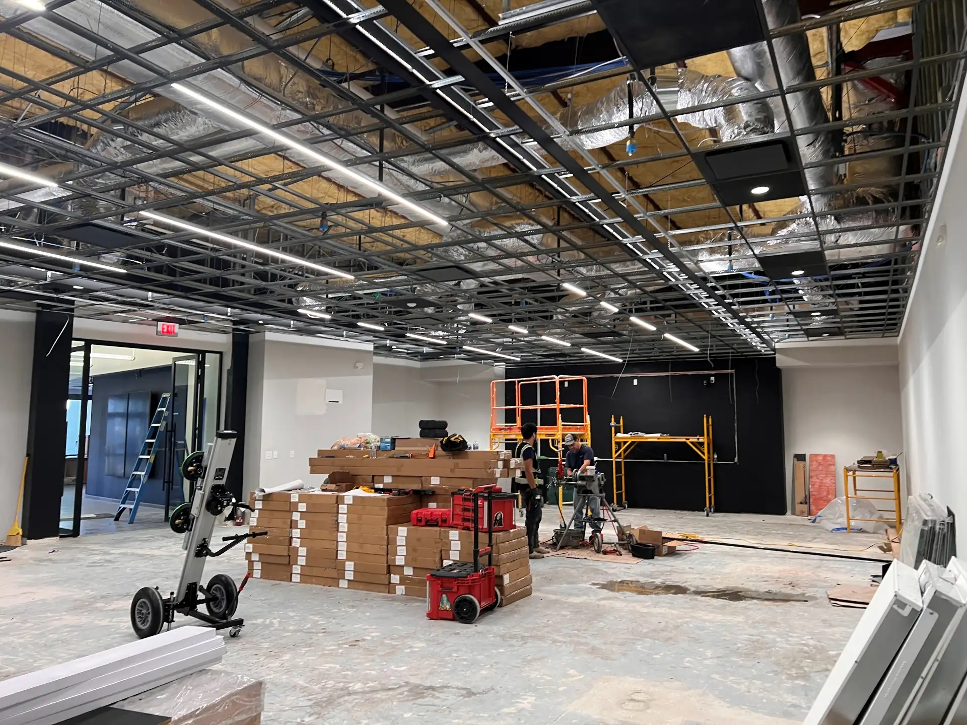A wide view of a large commercial room undergoing interior construction, showing workers, stacked materials, scaffolding, and an exposed ceiling grid with strip lighting installed.