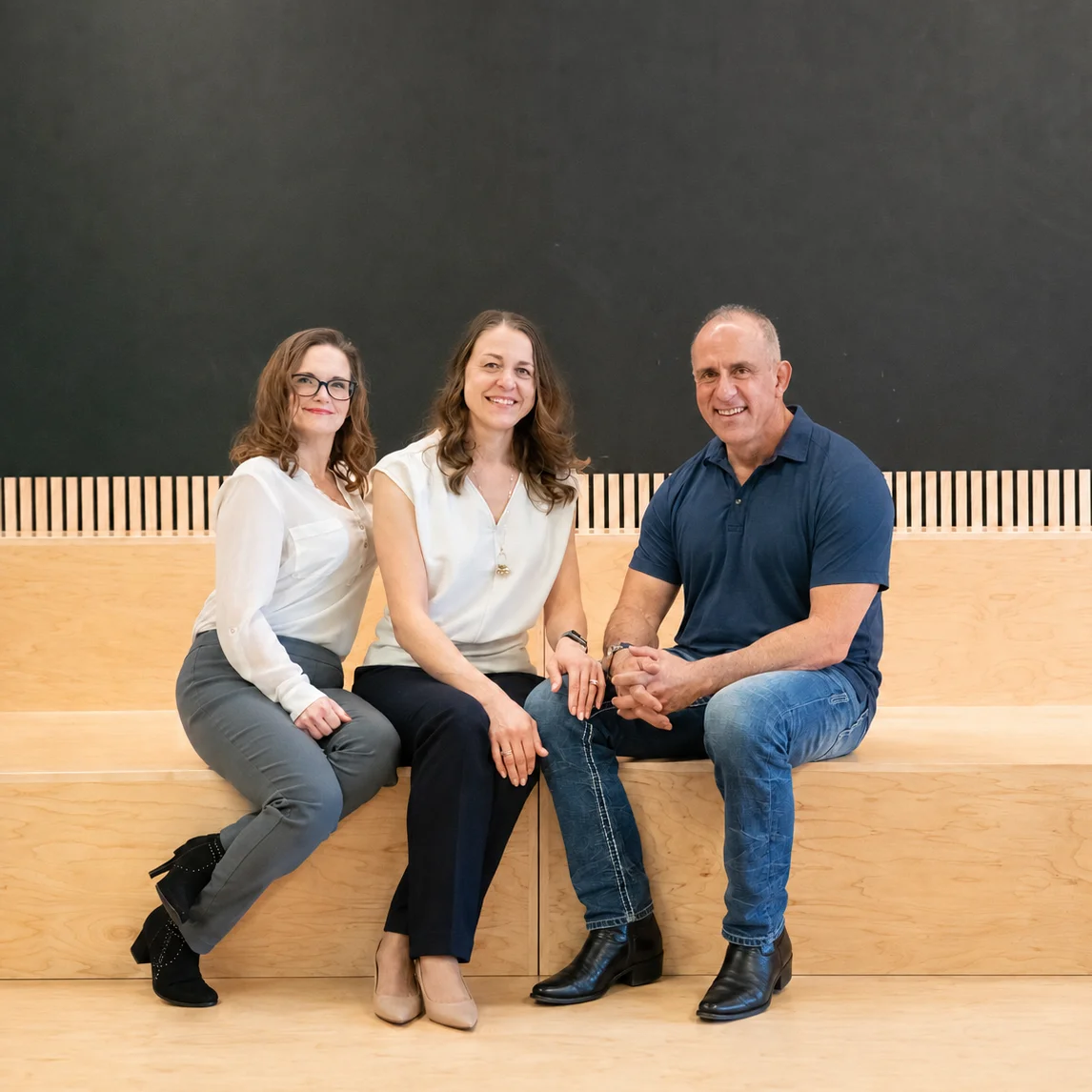 Three corporate leaders—two women and one man—sitting together on a tiered wooden bench against a dark background, smiling at the camera.