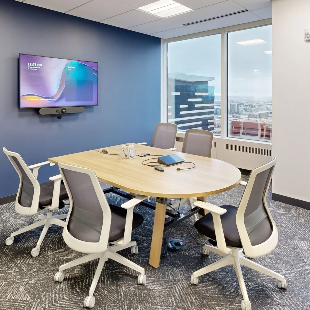 A modern small meeting room featuring a light wood oval table with six white and gray ergonomic office chairs, a wall-mounted TV screen on a blue feature wall, and large windows overlooking a city skyline.