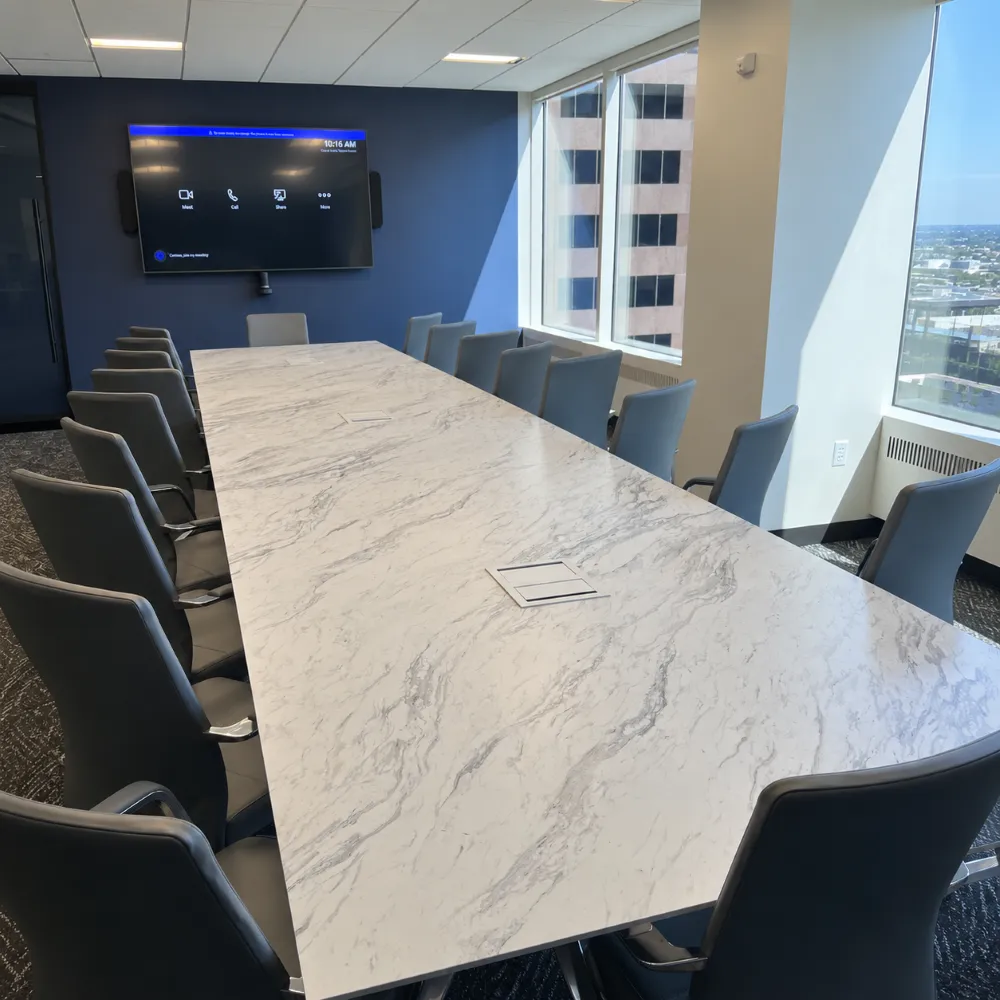 A high-angle view of a long, marble-patterned conference table with dark gray leather office chairs, located in a high-rise office with large windows overlooking a city skyline.