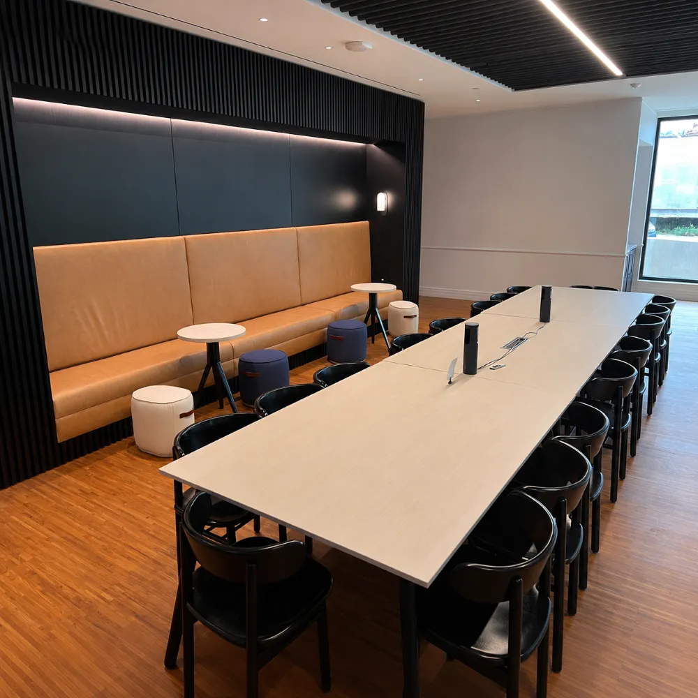 A modern breakroom featuring a long light-colored dining table lined with black chairs, opposite a recessed tan leather booth wall with small side tables and round ottomans.
