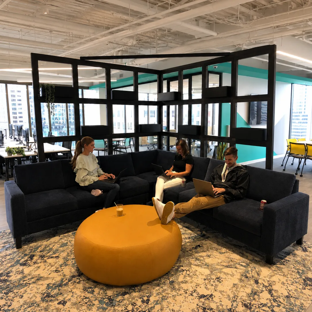 Three colleagues sitting with laptops on a dark blue sectional sofa around a large round yellow ottoman in a spacious, modern open-plan office.