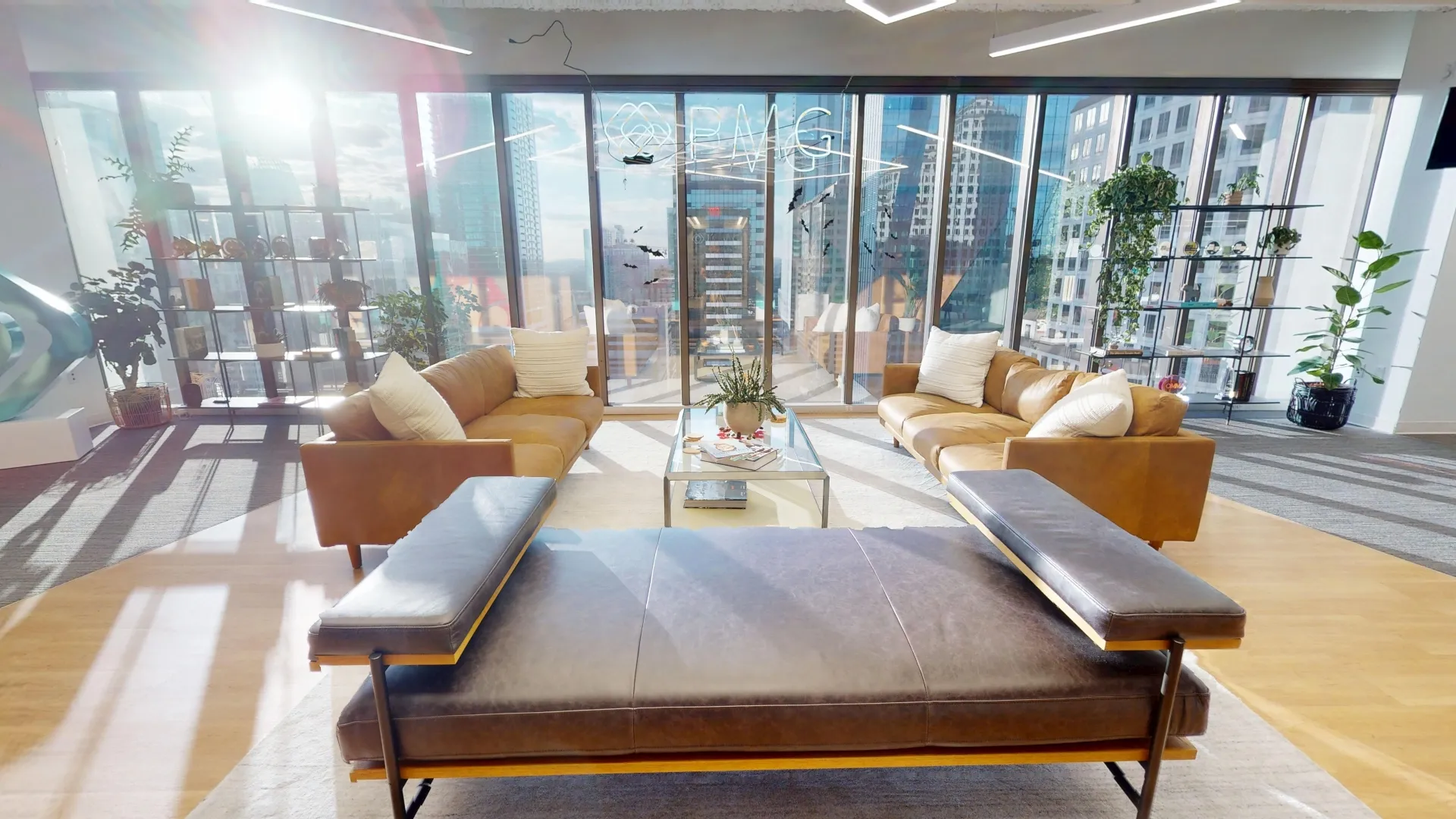 A sunlit executive lounge featuring tan leather sofas, a glass coffee table, and a dark leather daybed, set against floor-to-ceiling windows with a skyline view and a "PMG" glass sign.