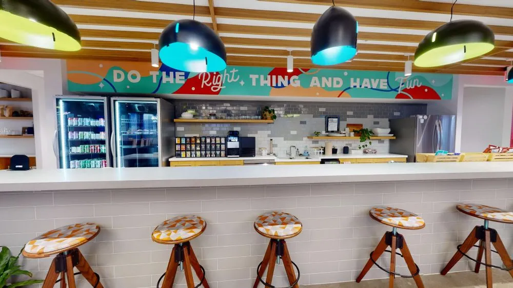 An office kitchen and bar counter featuring patterned wooden bar stools, grey tile backsplashes, and overhead pendant lamps beneath a colorful mural reading "DO THE RIGHT THING AND HAVE FUN".