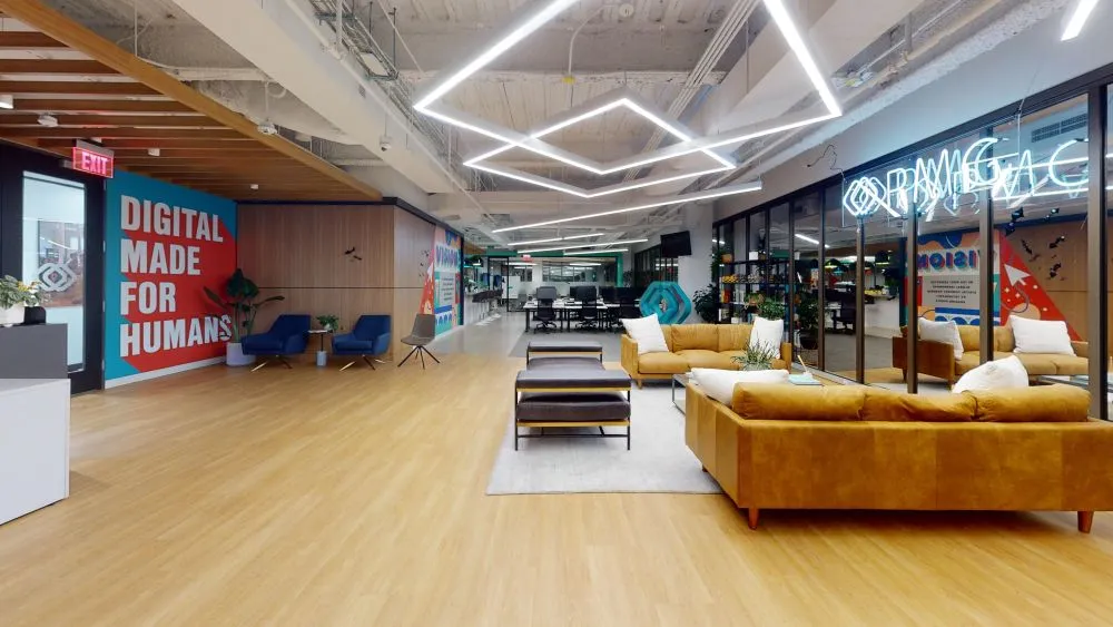A bright office lounge area with light wood floors, diamond-shaped ceiling light fixtures, tan leather couches, and a vibrant wall sign reading "DIGITAL MADE FOR HUMANS".