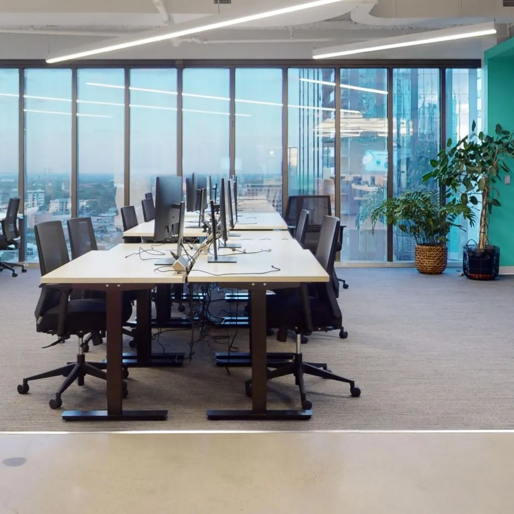 A row of modern office desks with computer monitors and black mesh swivel chairs, positioned in front of floor-to-ceiling windows overlooking a city view.
