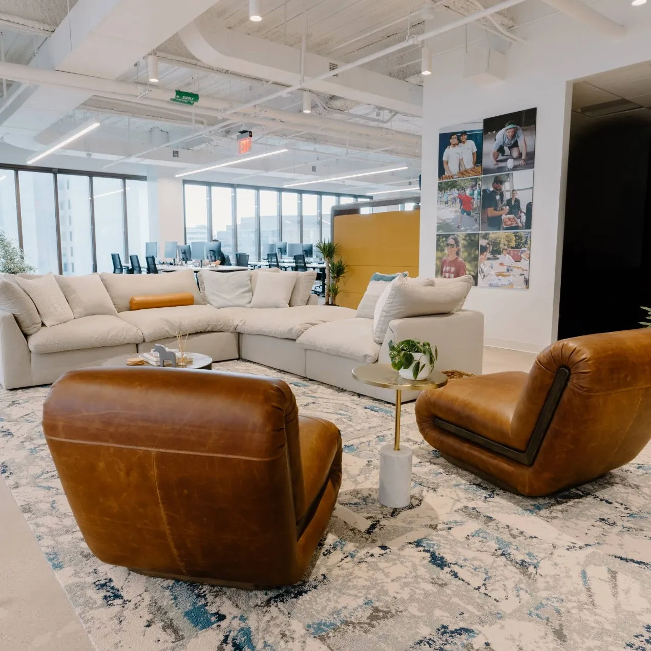 An open office lounge area styled with a large light gray sectional couch, two low-slung brown leather armchairs, and a blue-patterned rug.